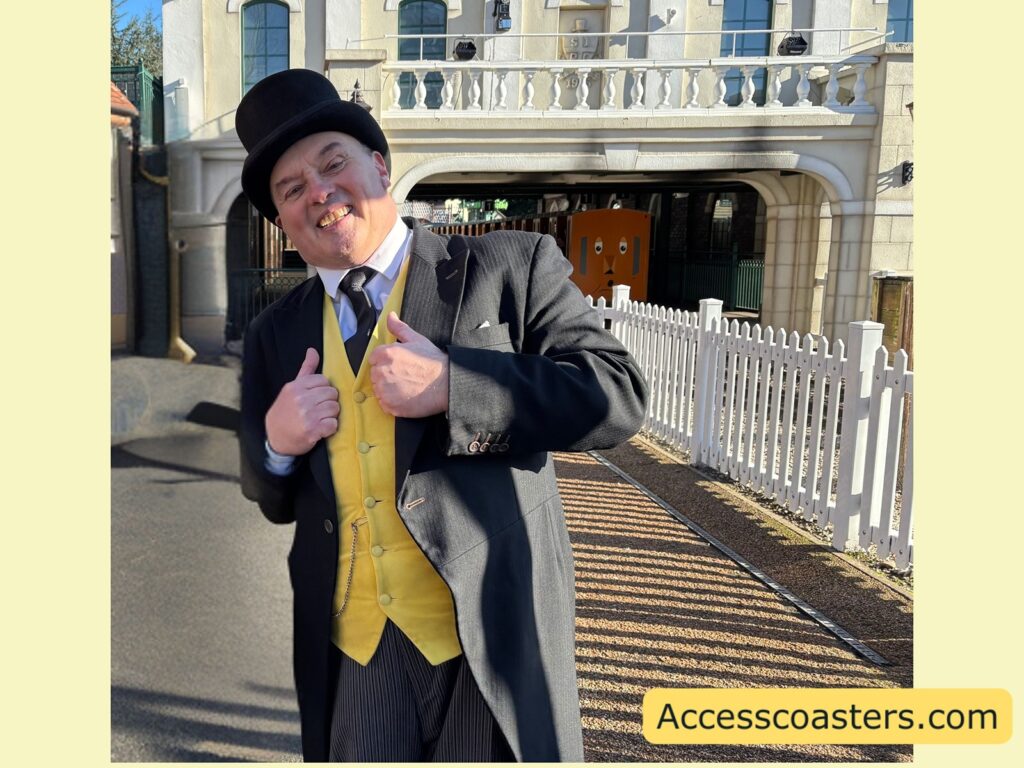 Sir Topham Hatt performer in a black top hat, black coat, and yellow vest poses and smiles beside a white picket fence in front of the Thomas Land entrance building and balcony.