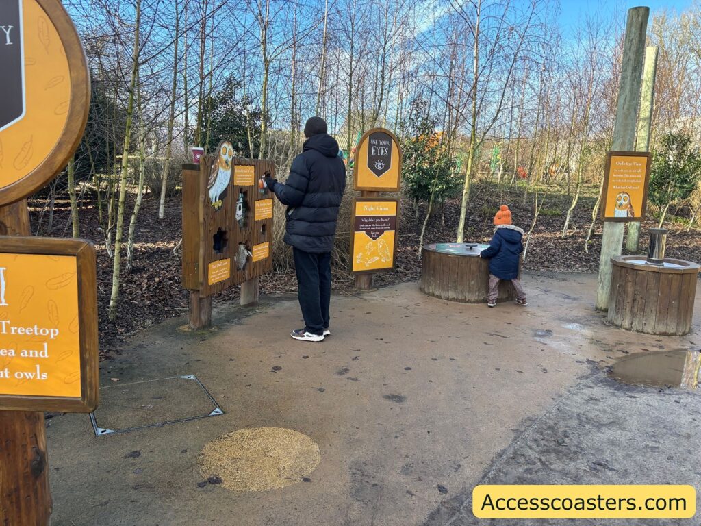 Outdoor activity station area with multiple owl-and-fox themed signs and hands-on posts, with visitors using the displays.