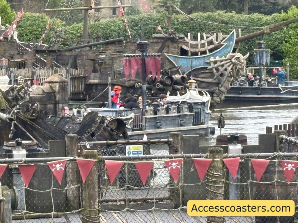 A close view of pirate-themed boats and dock structures packed with nautical props and netting; actors are on the boats, with red-and-white bunting along the fence in front and a pirate takeover overlay.
