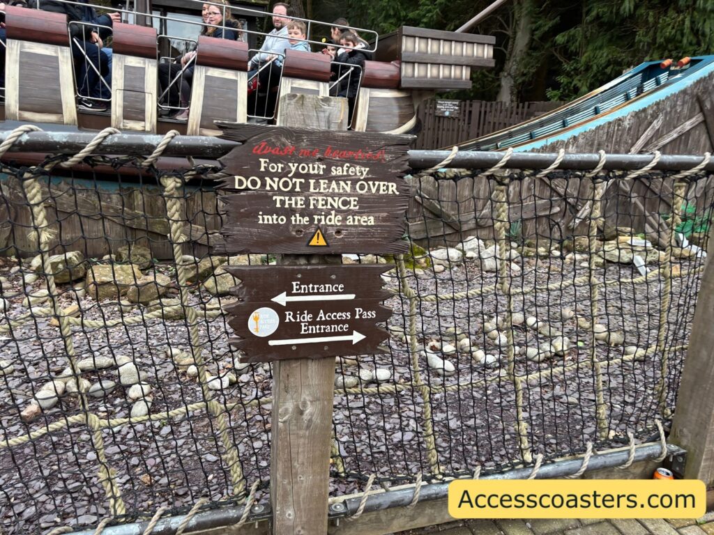 A netted fence in front of a rocky area, with a warning sign that reads: “For your safety DO NOT LEAN OVER THE FENCE into the Ride area” and a smaller sign below that reads “Ride Access Pass Entrance.”