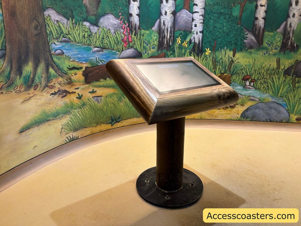 Interactive touch-table on a pedestal inside, positioned in front of a painted woodland mural.