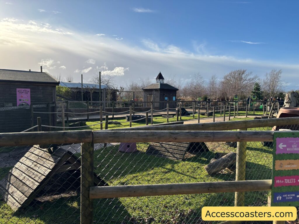 View across fenced goat walkthrough pens with wooden railings and small shelter structures under a bright sky.
