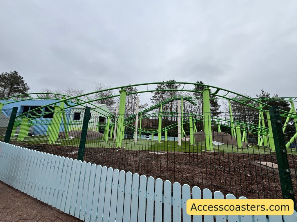 A green roller coaster track with multiple small hills and turns behind a pale blue picket fence.