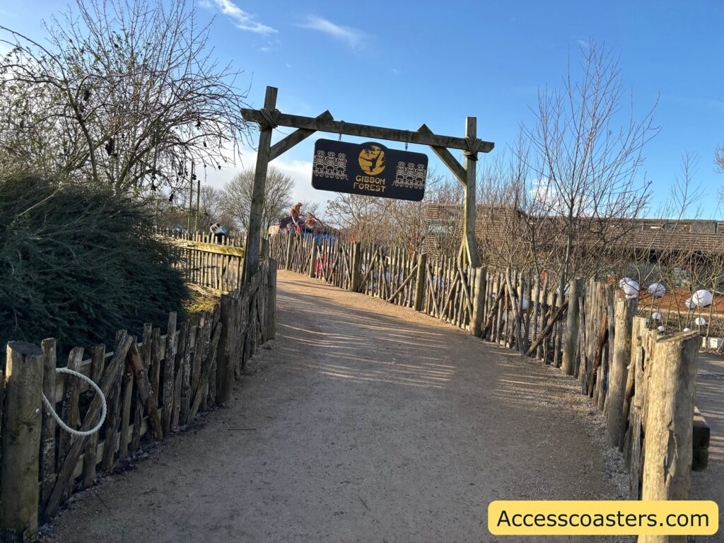 Outdoor entrance path to The Gibbon Forest, with rustic wooden fencing and a hanging sign over the walkway.

