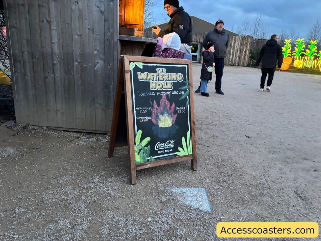 Marshmallow roasting area sign on a stand beside the path, with visitors and colorful lights in the background.

