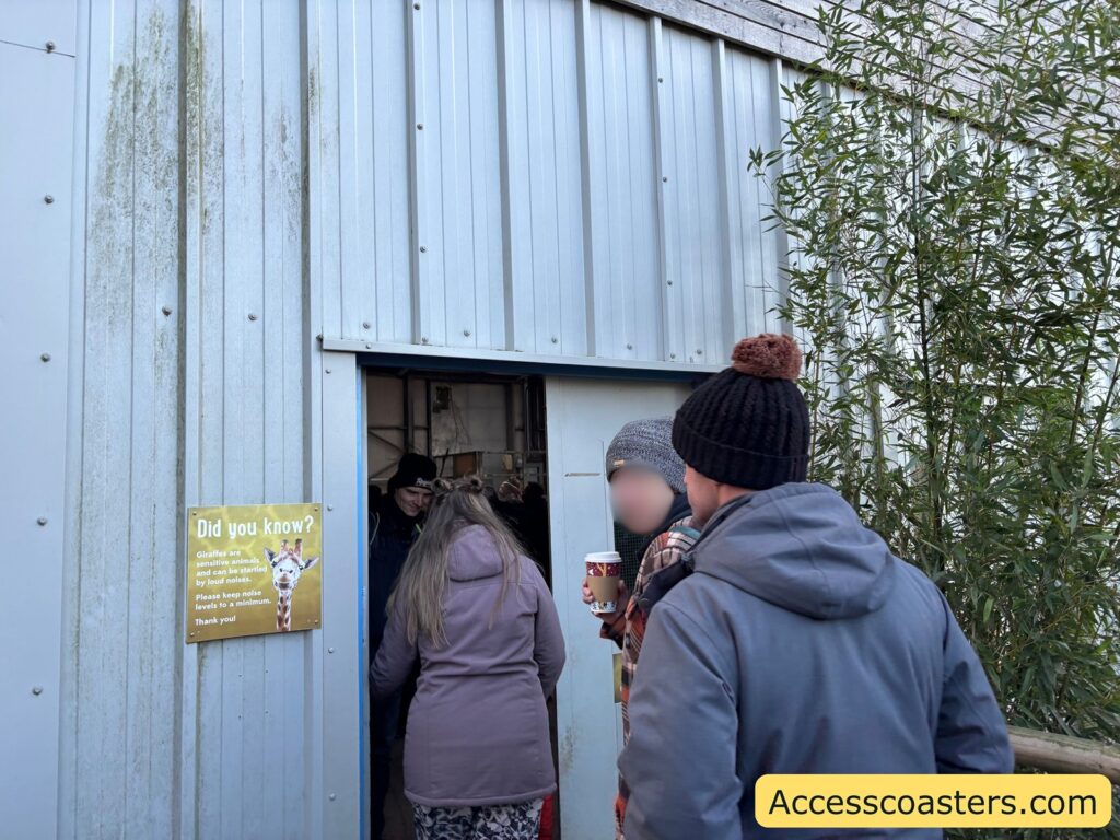 Visitors entering an indoor giraffe viewing building through a doorway in a metal-sided wall.
