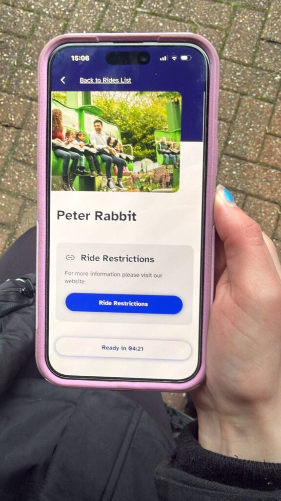 Smartphone showing a ride access pass app. The screen has a “Back to Rides List” header, a photo of riders on a green ride, the ride name “Peter Rabbit,” a “Ride Restrictions” section with a blue “Ride Restrictions” button, and a status line at the bottom indicating a ready time.