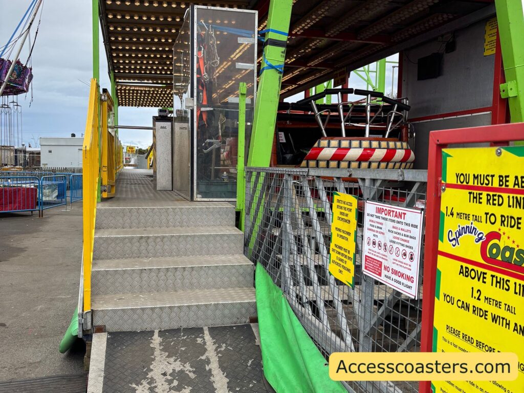 Entrance to the Spinning Coaster, showing a short flight of metal steps with handrails leading up to the platform, the side of the bright green track structure, and a section of the loading area with red and yellow ride cars waiting.
