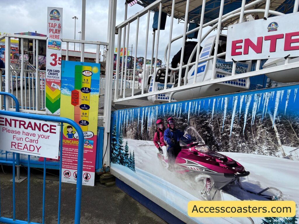 Matterhorn ride entrance with bright blue railings, multiple signs showing safety information and prices, and a snowy-themed mural of people riding snowmobiles along the base of the ride platform.
