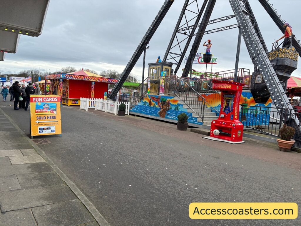 Large pirate‑ship ride structure on the left and a yellow A‑frame sign on the pavement pointing towards the arcade building where you carer tickets are. 