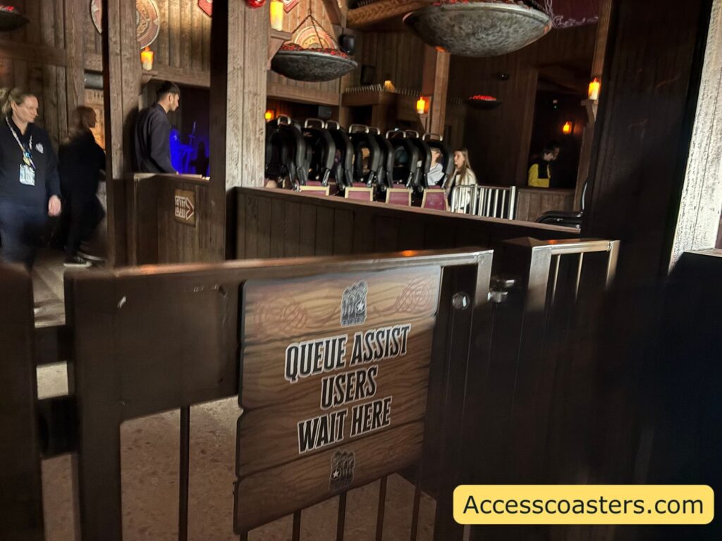 image of queue assist entrance gate to ride loading area with a sign that reads queue assist users wait here in the background you can see people getting on and off the ride with a train in the station. 