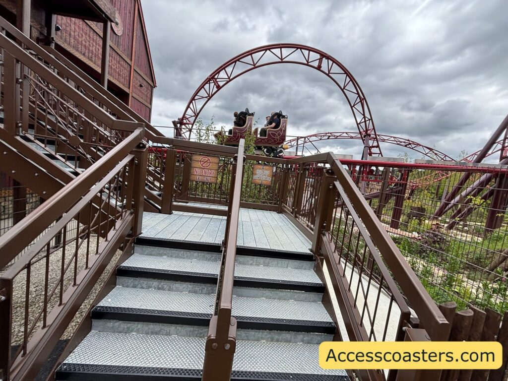 image of grey metal stairs with black edges and red handrail. in the background you can see a train on Drakon break run and the outside of the ride station. 