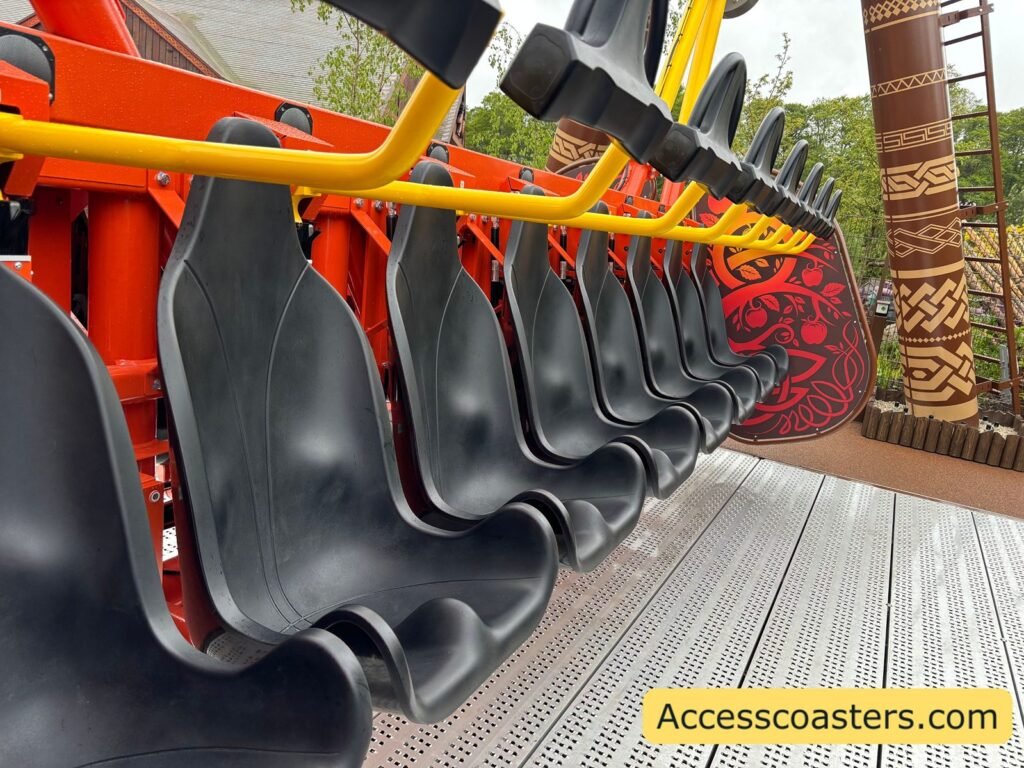 Close-up side view of Vild Swing’s bright orange and black individual seats in a row, each with its own lap bar, hanging from the ride arm above the platform.
