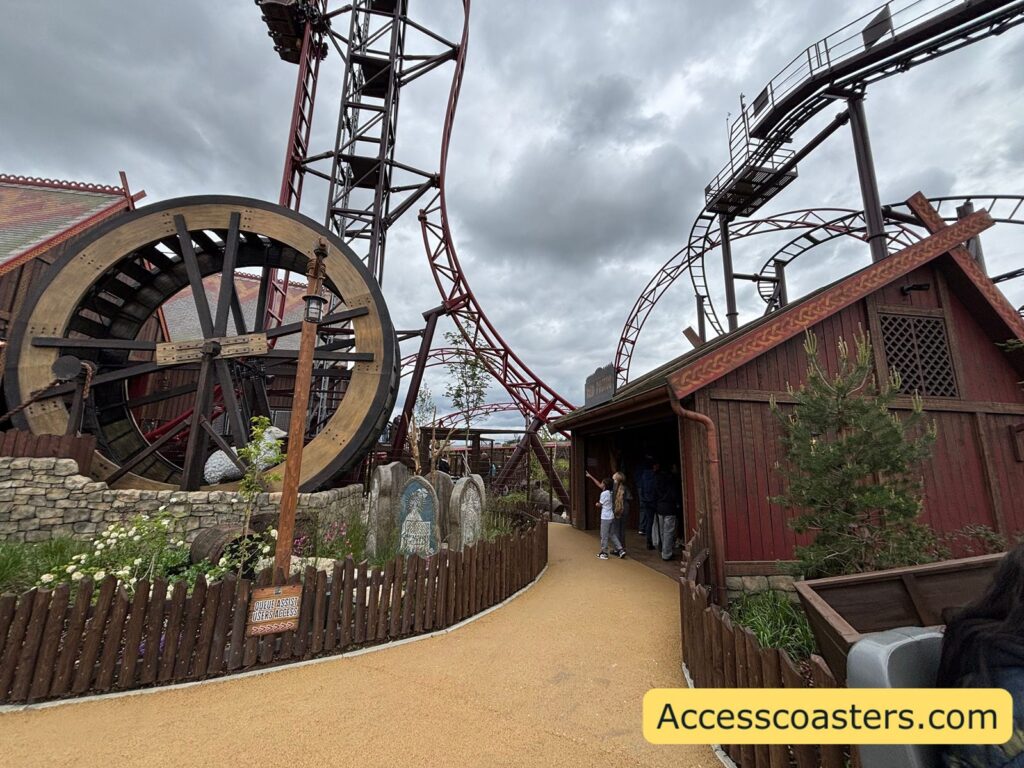 image of queue assist entrance to Drakon there is a sheep in a wheel at the side of the path as well as a queue assist entrance sign directing guests to the queue assist entrance on the otherside of the path there is a red wooded photo booth building. 