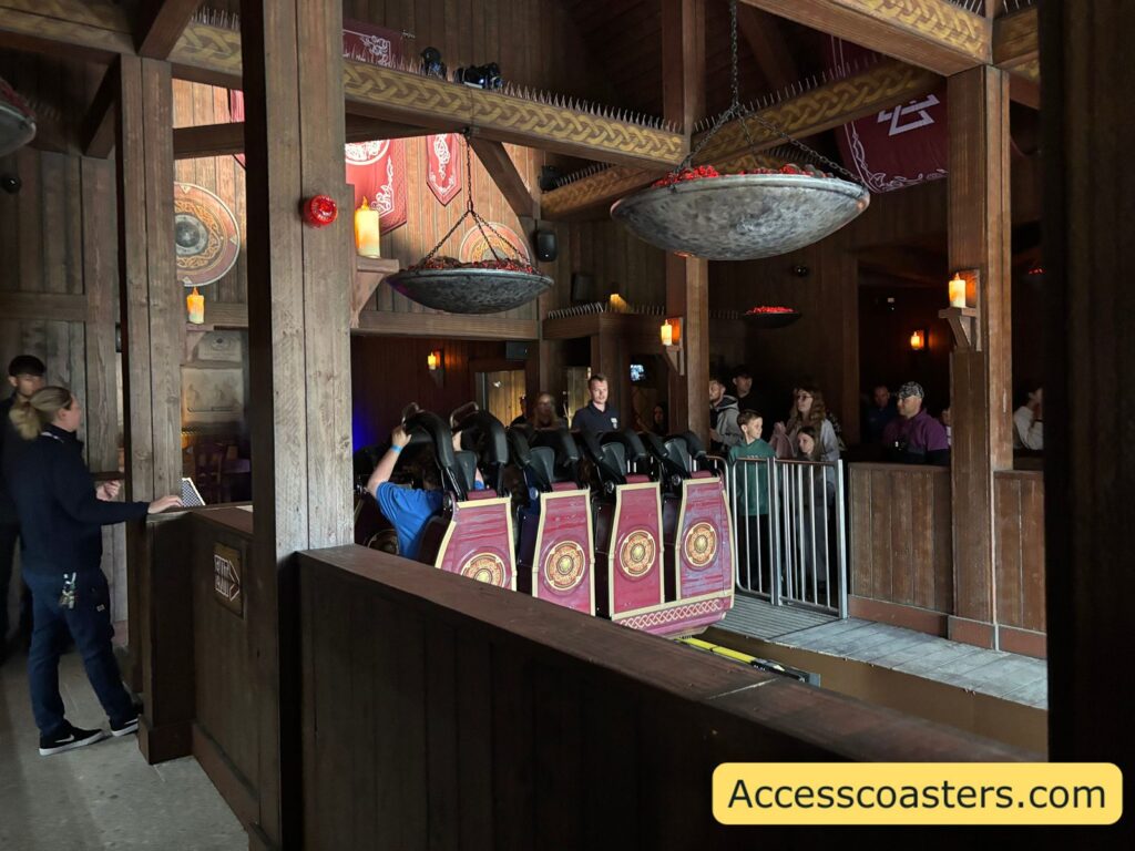 image of inside Drakon ride station there is silver air gates with people waiting for the ride the interior of the station is wooden with decretive shields and Viking banners on the walls. 