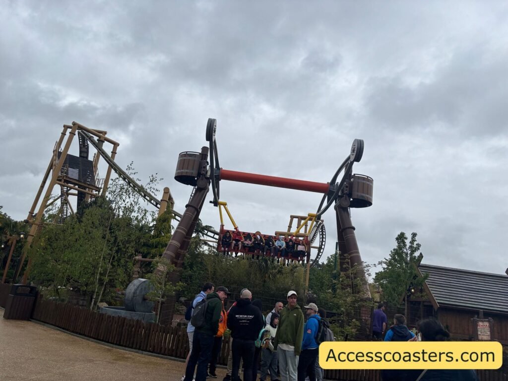 Wide shot of Vild Swing in motion on an overcast day, the large swinging arm tilted to one side with riders facing outward, surrounded by themed buildings and trees.
