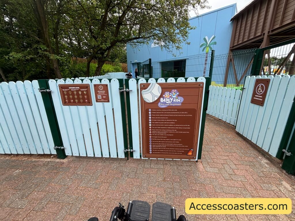 image of whelchair entrrance for bluey the ride there is a blue picket fence with an open gate with sign that reads wheelchair entrance leading to the wheelchair loading pont. the sensory level, restriction board and ride access pass check in point are all on the fence.
