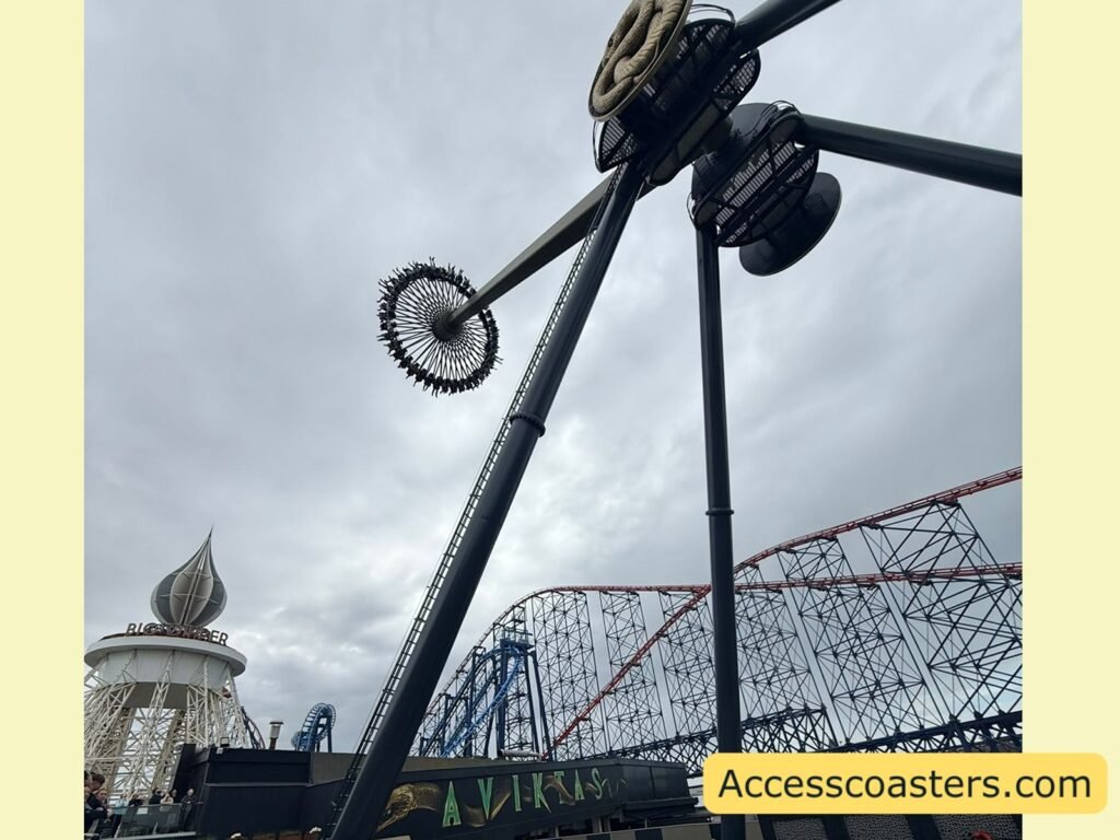 Upward view of Aviktas’ central hub and support towers against a cloudy sky, showing the circular ring of seats suspended below and portions of the surrounding roller coaster structure in the background.