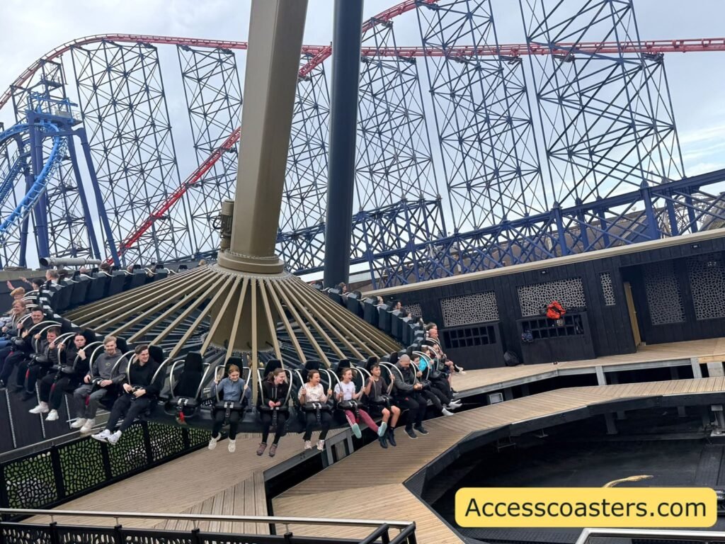 Wide view of Aviktas in motion at Blackpool Pleasure Beach, with riders swinging in a large circular gondola beneath tall black support arms, the Big One roller coaster’s red track and steel structure towering behind under a grey sky.
