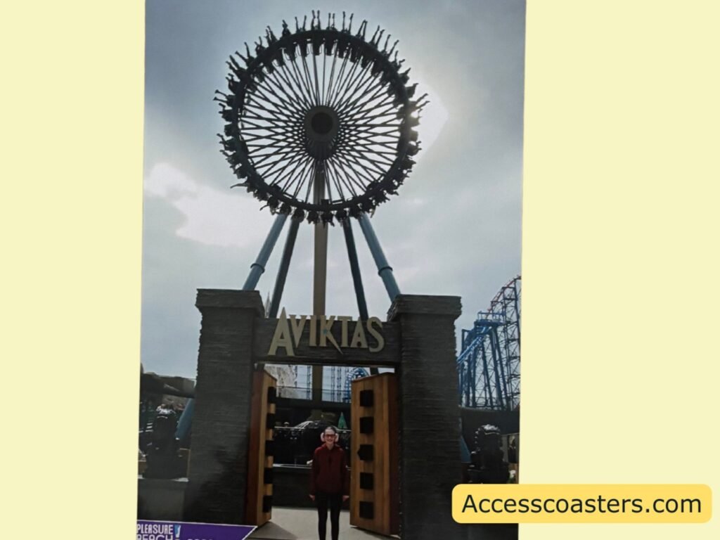 Portrait photo taken in front of the Aviktas ride entrance at a theme park. Two chunky grey stone-effect pillars frame open wooden doors, with a large circular spiked wheel structure towering above them. The word “AVIKTAS” is mounted across the top of the doorway, Iona is standing beneath the sign smiling