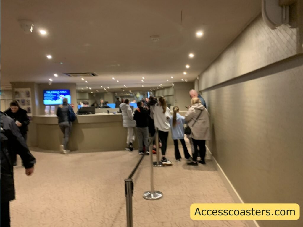 Indoor view of the Blackpool Pleasure Beach ticket office, showing a bright, modern lobby with a long counter at the back where several staff members are serving guests. A small queue of adults and children stands behind a short barrier leading up to the counter. The ceiling has rows of recessed lights and a TV screen on the left wall displaying information.