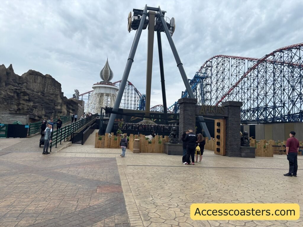 Tall grey A‑frame thrill ride “Aviktas” at Pleasure Beach Resort, with its swinging arm lowered over the boarding platform, people gathered in the paved plaza the big one track and big dipper ride building can be seen in the background.