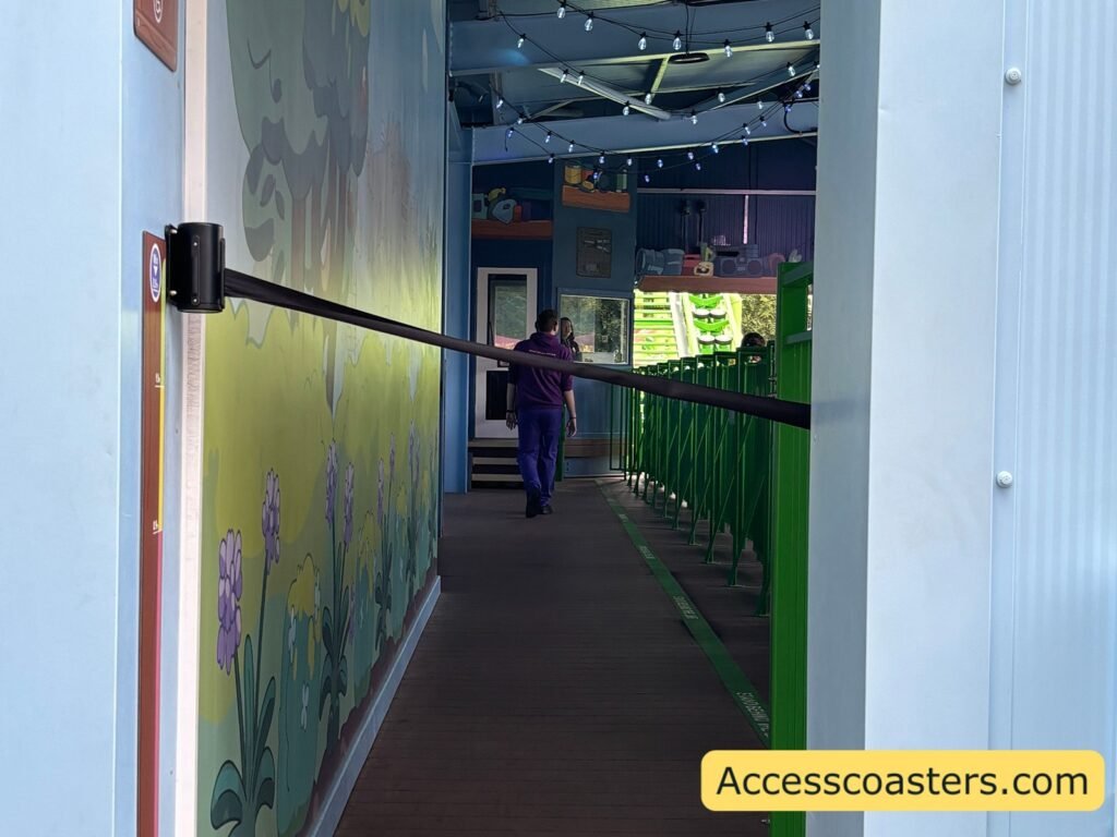 view of ride station from wheelchair entrance for bluey the ride there is a hight measurement just before entering the station a retractable barrier is across the entrance inside the station you can see green air gates and a garden themed painting on the wall as well as staff members waling about.