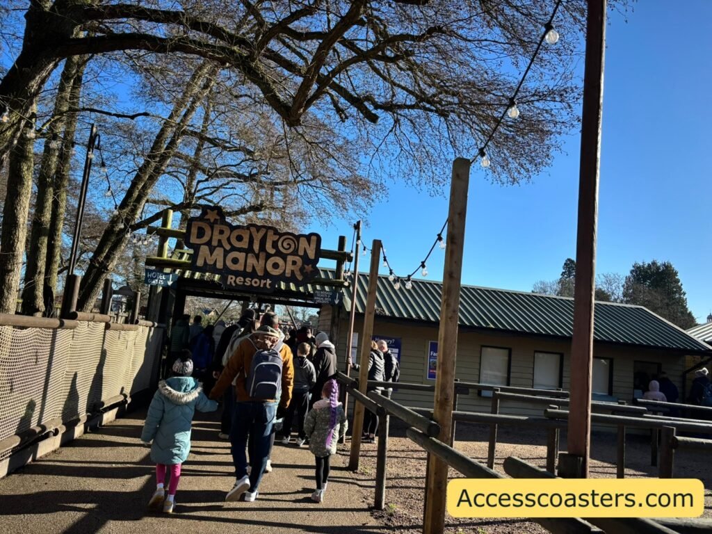 image shows wooden entrance to drayton manor there is a big wooden sign on top of the entrance gate of the drayton manor logo you can see the ticket office kiosks with a wooden fenced of queue to the right of the entrance and a queue of people at the entrance