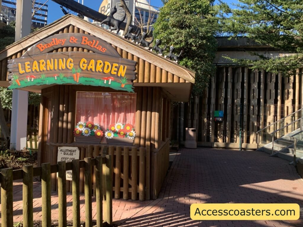 A small wooden building with a picket fence and a sign reading “LEARNING GARDEN” in bright letters, with cartoon-style artwork underneath. Trees and wooden posts are around the structure