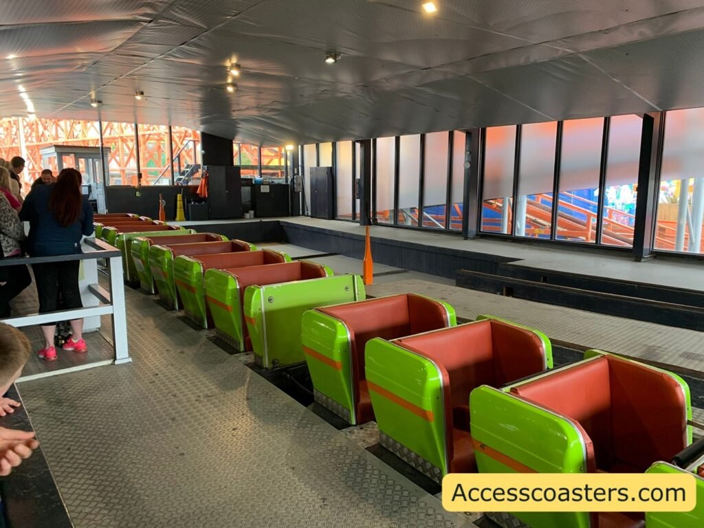 image of the ride station you can see people queuing and waiting behind the air gates as well as an empty train in the station. the train has orange seats and green ride carts  with orange stripes 