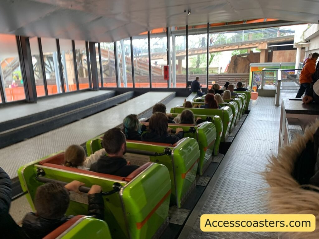 image shows station view from loading on to Nickelodeon Streak the train is full with 2 people sat per row staff members can be seen checking lap bards and people are waiting behind the queue line fences on the right. 