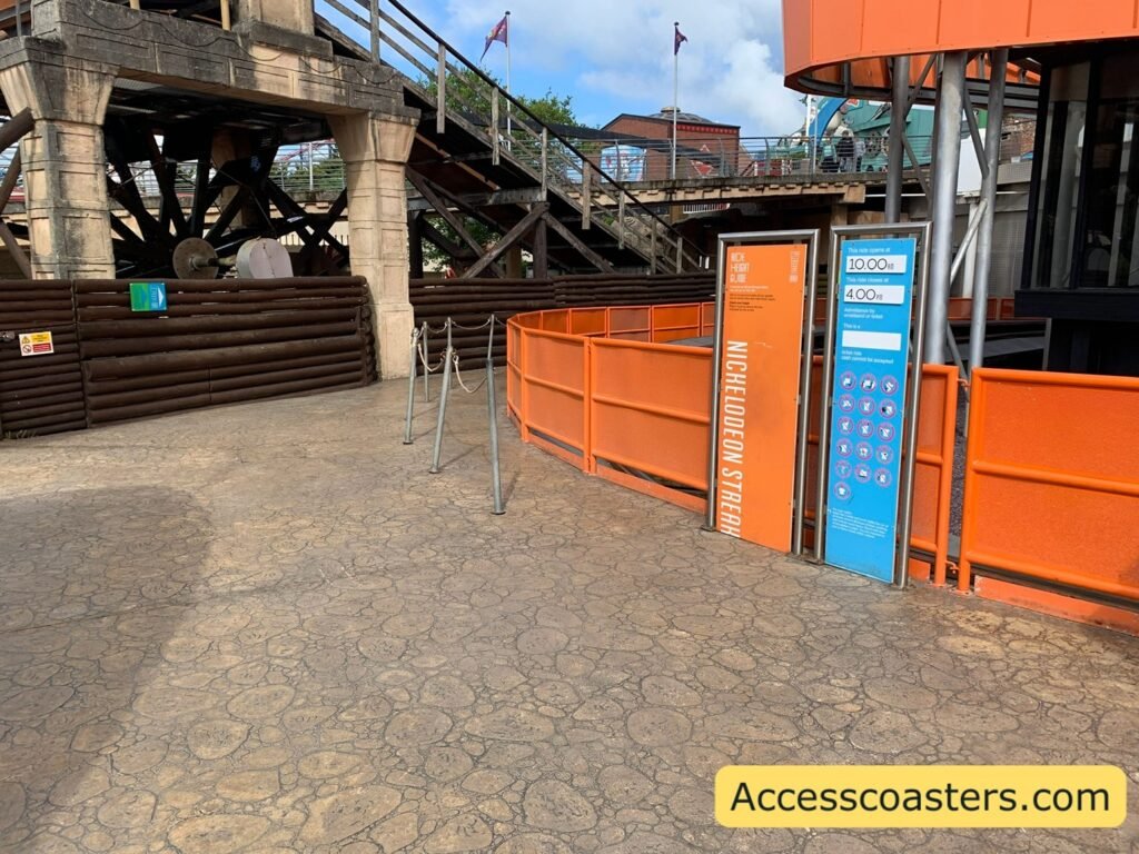 image shows a wide shot of the main entrance and easy access entrance for Nickelodeon Streak. there is the rides station on the right with the ride restriction signs that is the main entrance with a rope seperated the easy access entrsnce to the left that has sighs on the left wall that read easy access entrance and speedy pass entrance with arrows pointing down the queue line. 