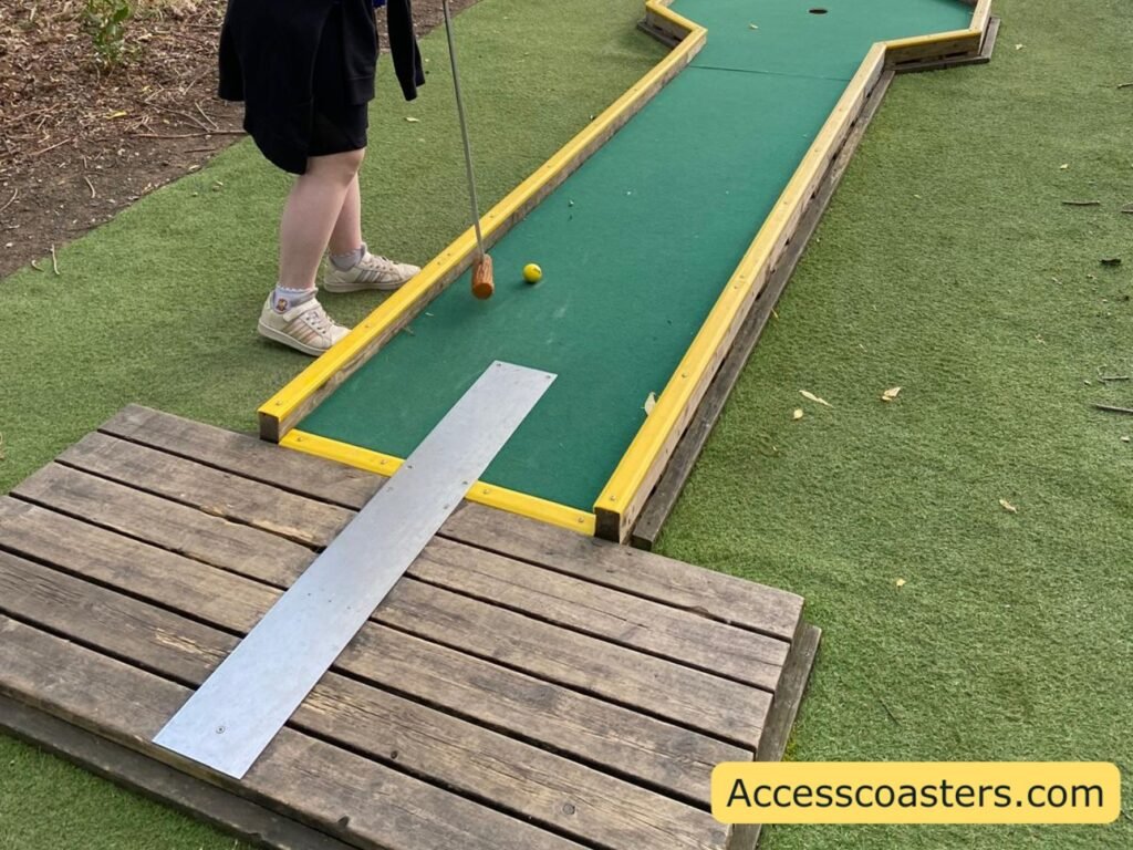 Person standing on artificial grass at the start of a Corn on the Club crazy golf hole, preparing to hit a yellow golf ball up a straight green fairway bordered with bright yellow edges, with a simple wooden ramp forming the tee area.
