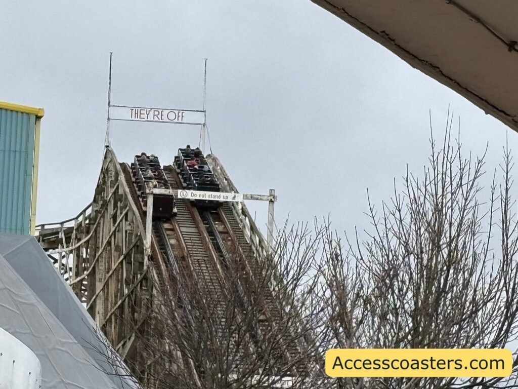 Exterior view of the Grand National wooden coaster lift hill with a train climbing, framed by bare trees and park buildings.
