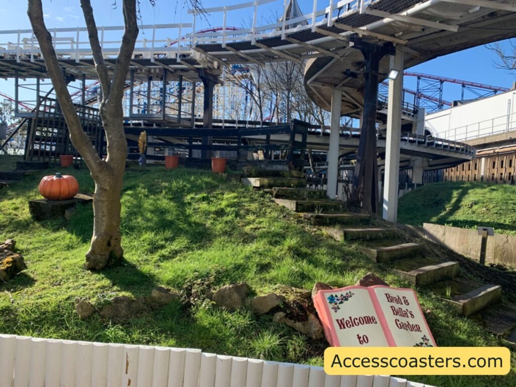 Grassy slope with a few bare trees and several large artificial pumpkins. Above and behind the hill is a steel rollercoaster track structure
