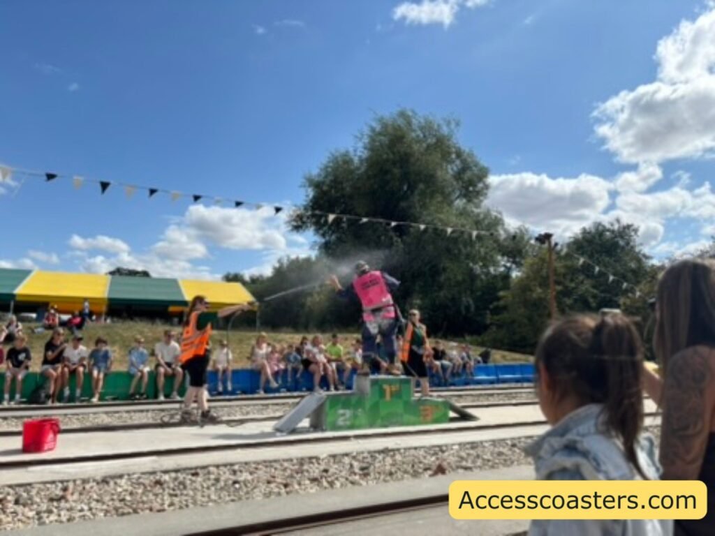 Action moment from the Cornula 1 show, taken from the side of the pebbled viewing area. Colourful track barriers cross the scene, and in the middle a performer in an orange outfit stands on a low green platform between the tracks while another performer in a bright pink top is dramatically lifted into the air on a water‑powered jet board, spraying water beneath them