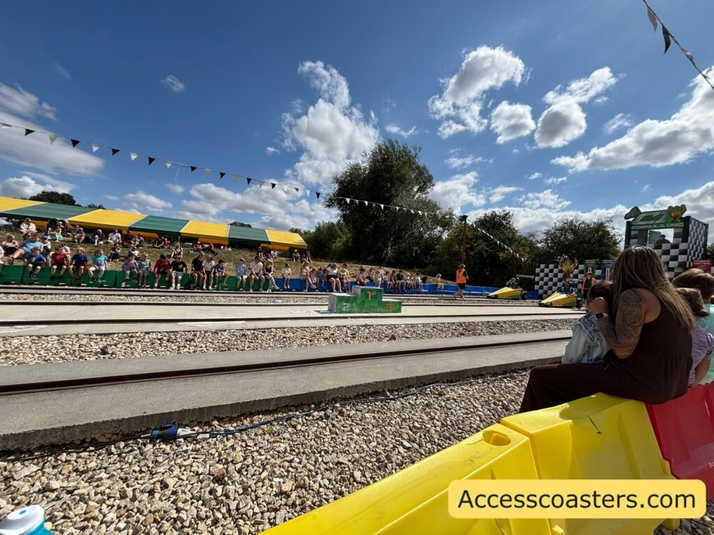 Cornula 1 show in progress on a sunny day, viewed from the side of the track. The parallel tracks and colourful barriers run across the foreground, while a large crowd of families watches from sloping grass banks and from long rows of basic bench seating under yellow‑and‑green striped awnings. 