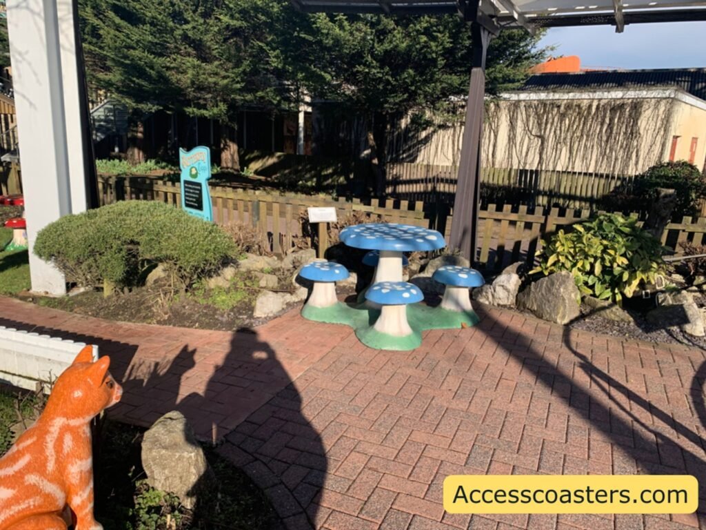 A paved brick area with low, mushroom-shaped stools arranged like a children’s seating area, next to bushes and a fence. There is a decorative animal figure 