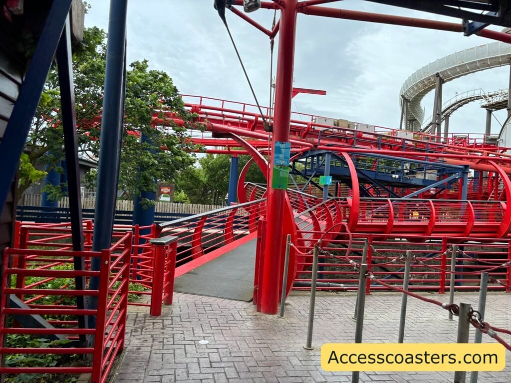 Closer view of the same outdoor area for wheelchair and easy access, with guests moving under the red coaster track and steel canopy toward the entrance point with signs reading easy access pass entrance and a ramp leading to the exit