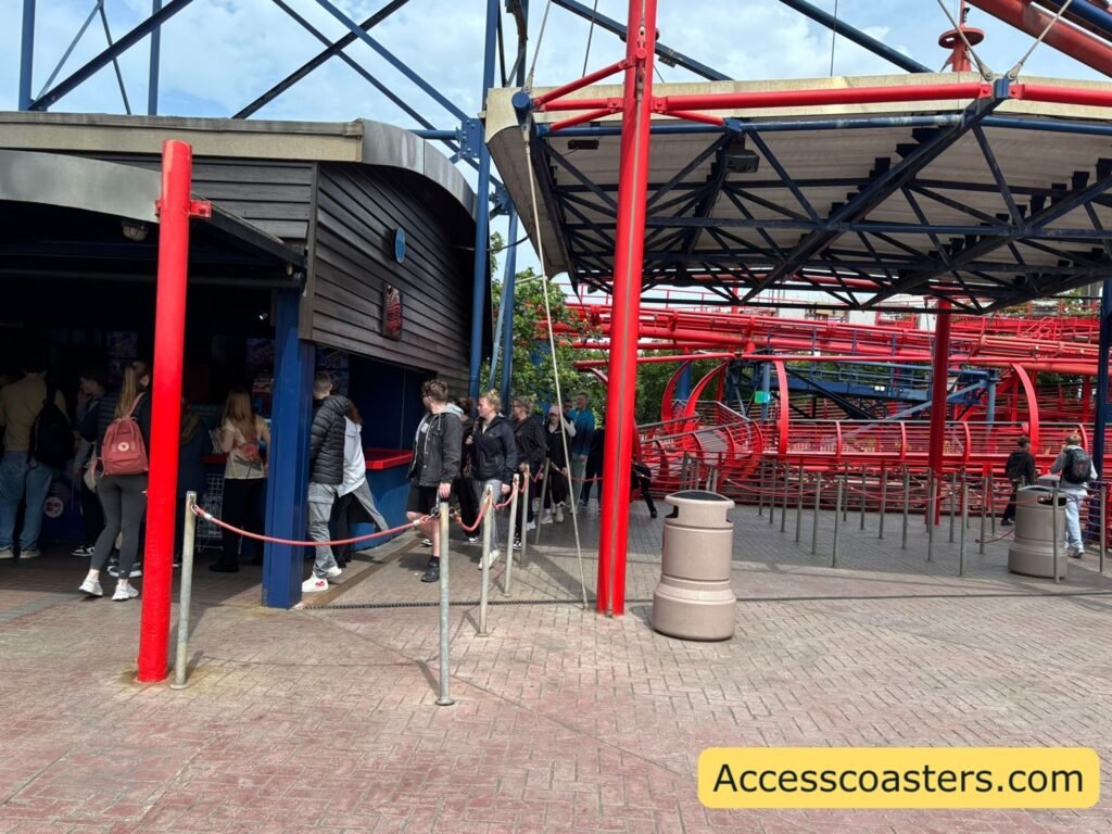 Outdoor view of the Big One’s easy access entrance beside the main station building, with red railings and steel coaster supports overhead, showing the route toward the ride exit and photo booth area.