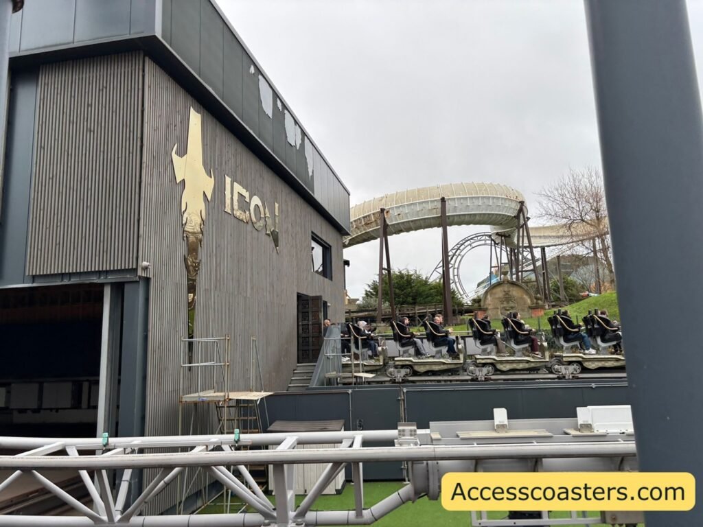 The main ICON station building with a modern, boxy design and the word “ICON” with a bird-shaped logo on the wall. Track runs in front of the building, and a coaster train with riders is visible to the right.