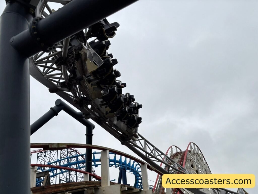 A closer view under the coaster where a train full of riders is inverted on a twist in the track, supported by black steel beams. Another part of the coaster and surrounding park structures are in the background.