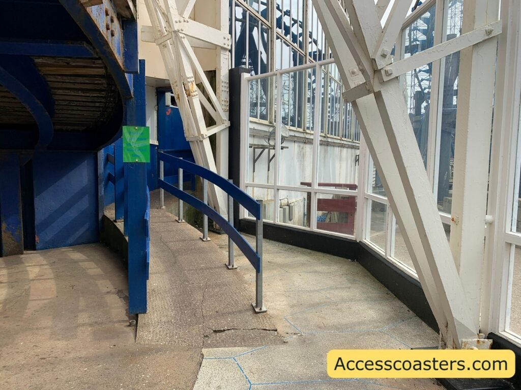 image of ride exit/easy access entrance to big dipper. you can see a rap with blue railings and a green sign with text that reads  please wait here to be called. 