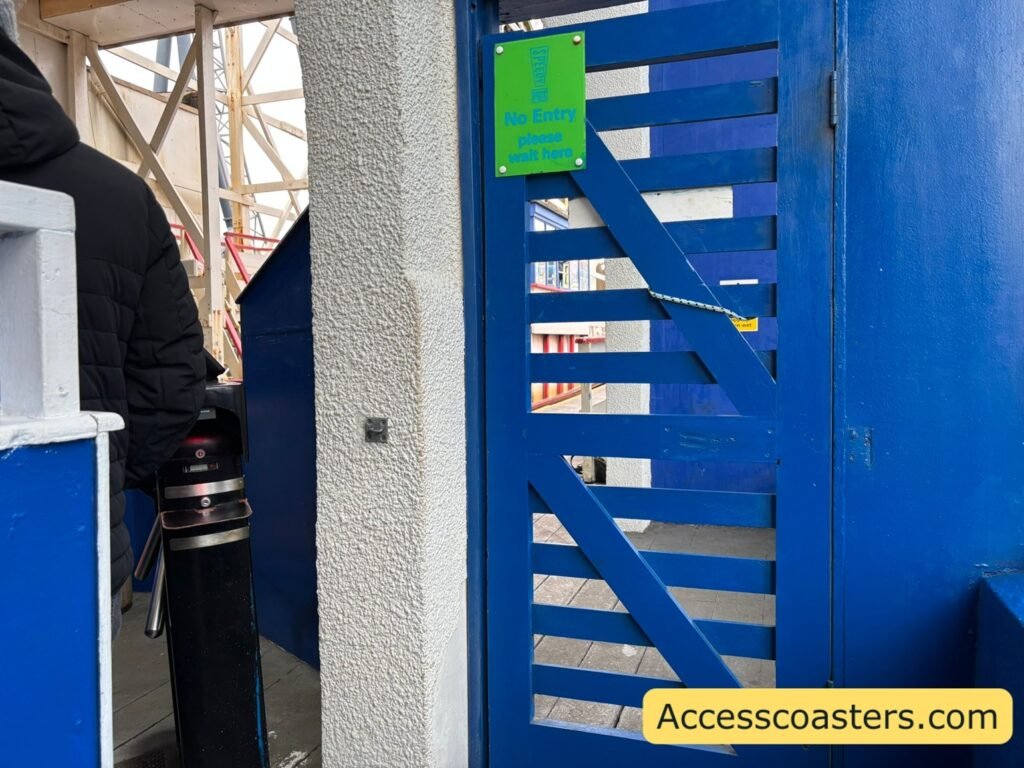 image if front of easy access queue line you can see a divider with people waiting in the main queue on the other side, a turnstile and a blue gate with a sign that reads no entry please wait here. 