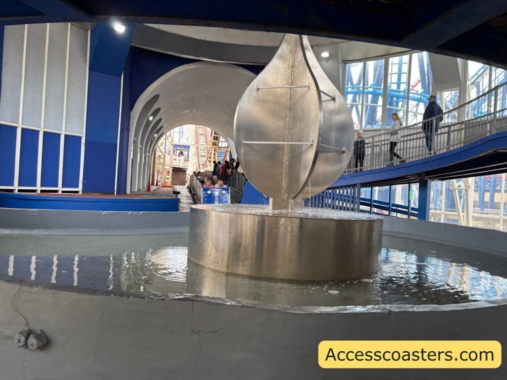 image of big dipper station and queue line you can see a big cercal water feature in the background the big dipper train is loading in the station  and you can see people queueing to the right. 