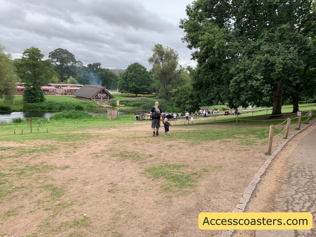Falconers quest show area from the level area  you can see grassy hill where audiences sit, the dirt and stone path along the right, the river at the bottom of the slope and, across the water, the wooden show building and a passing red-and-cream train surrounded by trees.