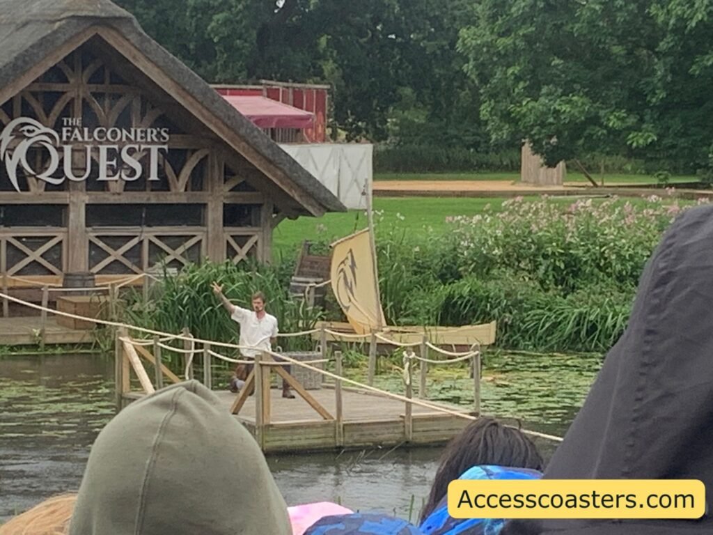 Closer view of The Falconer’s Quest wooden show building on the river with the large sign on the front, “The Falconer’s Quest”, and a presenter in a white shirt standing on a small wooden platform over the water, speaking to the audience 