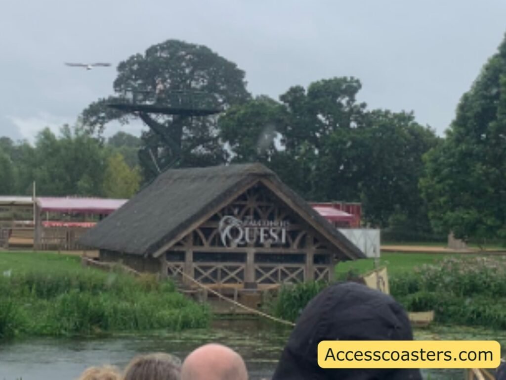 Side view of The Falconer’s Quest thatched wooden show building beside the water, with a large bird of prey gliding low across the sky above the trees and the tops of audience members’ heads. there is also a large Crain with a platform above the show building that the birds fly from