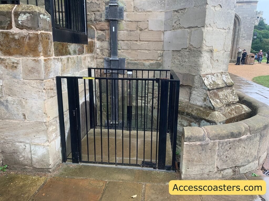 Stone castle entrance with a short flight of steps leading to a wooden door, blocked at the bottom by a black metal wheelchair lift platform and railings, showing the accessible side entrance in front of The Kingmaker.
