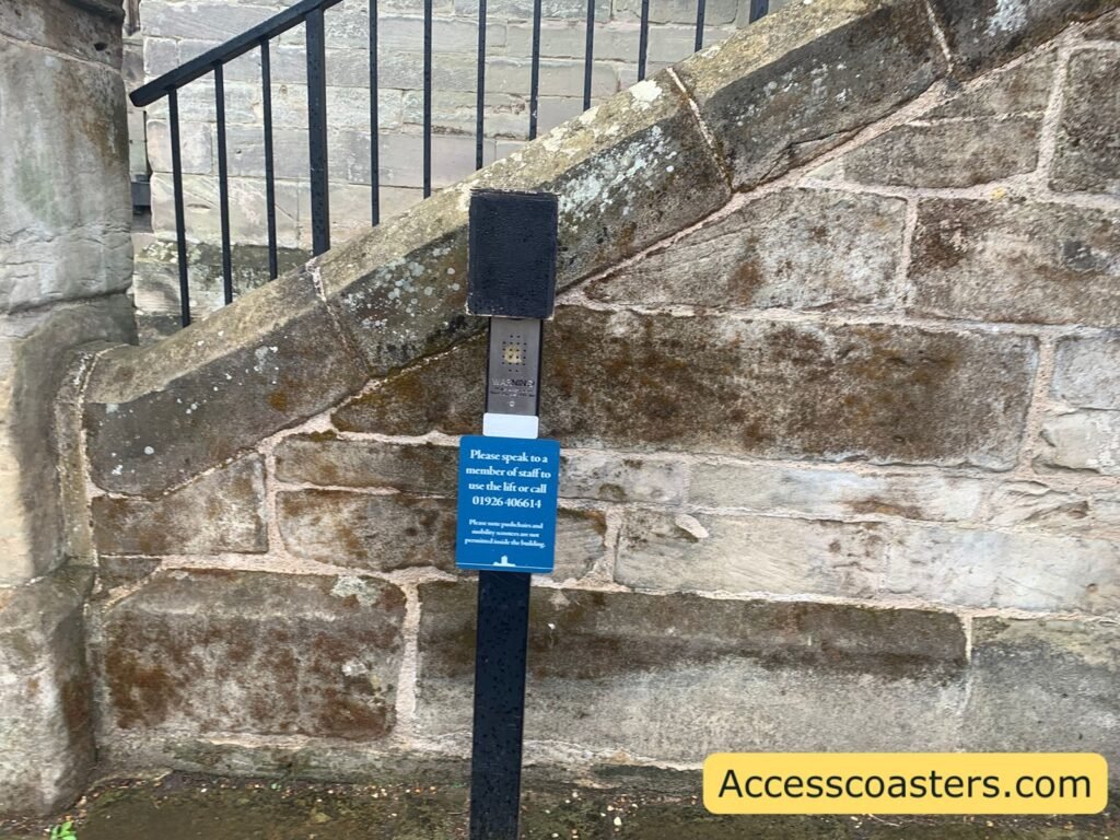 Close-up of the stone wall and steps beside the castle’s side entrance, with a blue information sign mounted next to the wheelchair lift controls, explaining how to call staff to operate the lift.